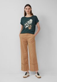 T-shirt à manches courtes vert foncé avec imprimé floral métallique, associé à un pantalon large beige clair avec ourlet replié et des baskets blanches.