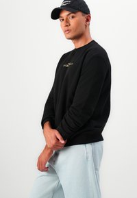 Sweatshirt preto com um logótipo dourado, gola redonda e mangas longas, combinado com jeans de ganga azul claro. Textura suave.