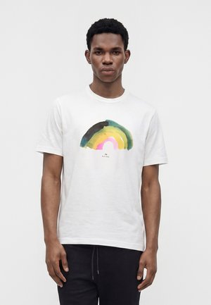Jeune homme debout, portant un t-shirt blanc avec un motif abstrait multicolore arc-en-ciel et un pantalon noir sur fond blanc uni.
