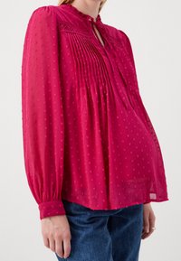 Blouse fuchsia à manches longues, détails floraux brodés, ouverture en goutte d'eau au col, et design plissé sur le devant, associée à un jean bleu.