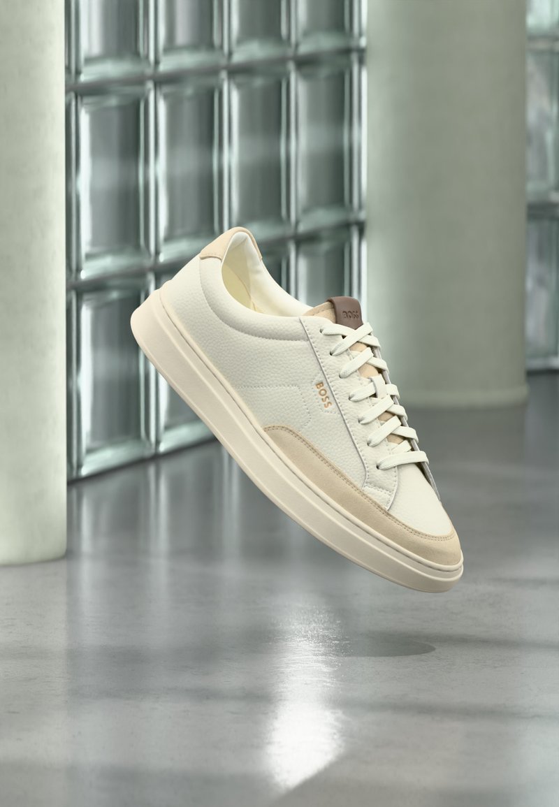 Άσπρο sneaker χαμηλού τύπου με μπεζ λεπτομέρειες και κορδόνια, με το λογότυπο "BOSS", αιωρείται πάνω από ένα γυαλισμένο δάπεδο με φόντο έναν τοίχο από γυάλινα τετράγωνα.