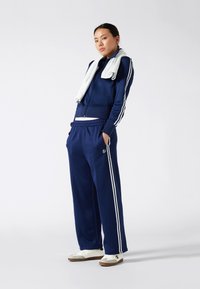 Marineblauwe tracksuit van zacht materiaal met witte strepen, jasje met ritssluiting, broek met elastische tailleband en witte sneakers met bruine zolen.