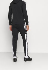 Schwarzer Hoodie und passende Sweatpants mit weißen Seitenstreifen, ausgestattet mit verstellbarer Kapuze und gerippten Bündchen. Schwarze Fußballschuhe vervollständigen das Outfit.