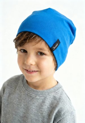 Young child smiling, wearing a bright blue beanie with a black label, and a light gray sweatshirt against a plain white background.