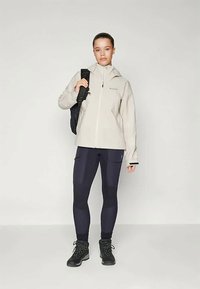 Giacca zip-up grigio chiaro, leggings blu navy e scarponi da trekking neri. Zaino appeso sulla spalla sinistra. Focus su abbigliamento sportivo e utilità.