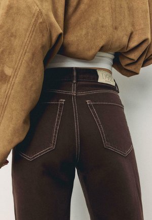 Osoba u tamnosmeđim trapericama s bijelim šavovima i svijetlosmeđoj kratkoj jakni, prikazana od struka prema dolje, okrenuta leđima.