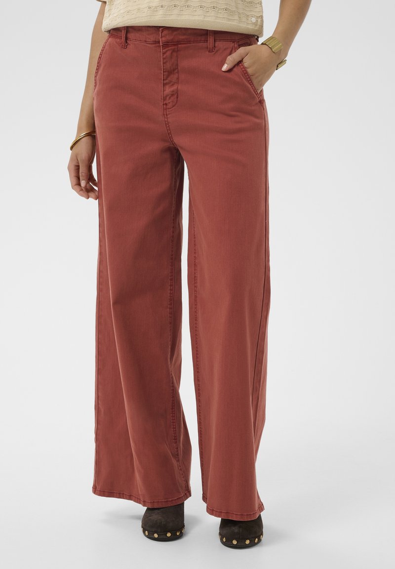 Personne portant un pantalon ample couleur rouille avec poches, un haut en maille beige, des chaussures marron cloutées, un bracelet en or et une montre sur fond blanc.