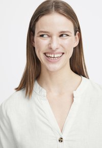Chemise en lin de couleur claire avec un décolleté en V, une texture subtile et un accent de bouton circulaire près du col. Visage souriant avec des cheveux raides.