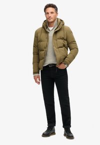 Chaqueta de plumas verde oliva con capucha, cierre frontal con cremallera, puños de canalé, llevada sobre un suéter de punto gris claro y unos jeans negros. Botas negras con cordones.