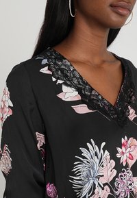 Blouse noire à fleurs avec des fleurs roses et bleues, un décolleté en V avec une bordure en dentelle noire, et une texture de tissu lisse et fluide.