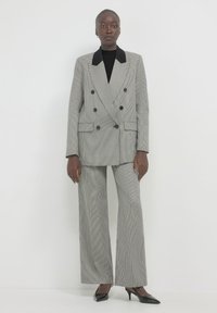 Donna che indossa un dolcevita nero, un blazer grigio a scacchi doppiopetto con colletto nero, pantaloni larghi abbinati e tacchi a punta neri.
