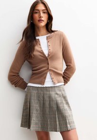Cárdigan de canalé en tono marrón con botones, superpuesto a una blusa blanca; combinado con una falda de cuadros plisada en tonos beige y gris, con líneas sutiles.