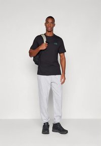 Zwart T-shirt met een klein blauwgroen logo, grijze joggingbroek en zwarte sportschoenen. De persoon draagt een zwarte rugzak.