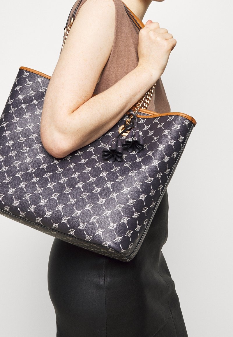Femme portant un haut marron sans manches, tenant un sac à main noir à motifs avec des anses en chaîne dorée et des breloques à pompons sur l'épaule.