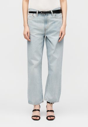 Person iført lyseblå vidde jeans med sort bælte og sorte åbne tåhæle, stående mod en ensfarvet baggrund.