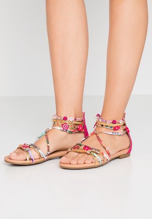 Pieds portant des sandales plates à bouts ouverts avec des brides multicolores, des accents dorés, et des bracelets de cheville en perles roses et fleurs sur un fond neutre.
