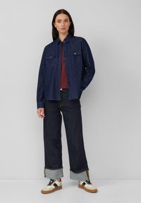 Dunkelblaue Denim-Hemd mit Knopfverschluss und zwei Brusttaschen, kombiniert mit weit geschnittenen dunkelblauen Jeans mit umgeschlagenen Säumen und lässigen Schuhen.
