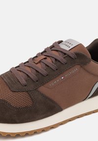 Sneaker marrone realizzato con materiali in rete e suede. Presenta lacci piatti, suola bianca e un'etichetta con il logo sulla linguetta, con accenti grigi sottili.