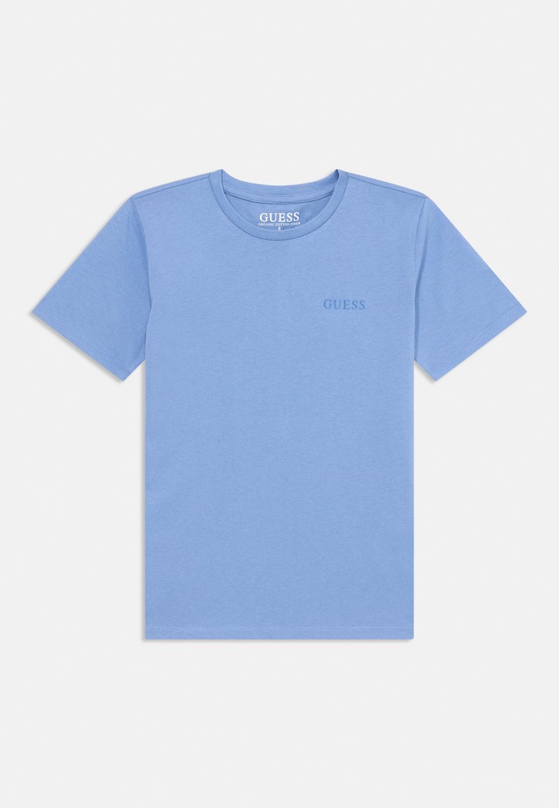 T-shirt à manches courtes bleu clair en coton bio. Présente un col rond et un logo "GUESS" discret imprimé près de la poitrine. Texture lisse.