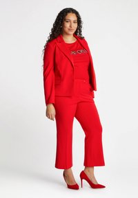 Czerwony garnitur skrojony na miarę z dopasowaną marynarką i skróconymi spodniami, w połączeniu z czerwonym topem z błyszcząca literą i czerwonymi szpilkami.