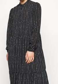 Černé šaty s knoflíkovým zapínáním vpředu, kulatým výstřihem, dlouhými rukávy a bílými vertikálními pruhy s vrstvenou sukní.