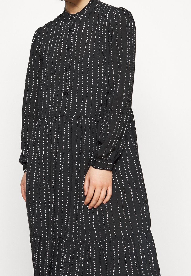 Černé šaty s knoflíkovým zapínáním vpředu, kulatým výstřihem, dlouhými rukávy a bílými vertikálními pruhy s vrstvenou sukní.