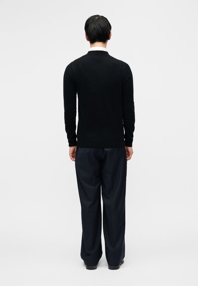 Μαύρο πουλόβερ με εφαρμοστή σχεδίαση, συνδυασμένο με σκούρο παντελόνι με ρίγες. Το υλικό φαίνεται λείο· το πουλόβερ έχει στρογγυλή λαιμόκοψη.