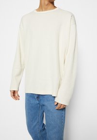 Person klädd i en löst sittande, långärmad ribbad krämfärgad tröja med rund hals, matchad med blå denimjeans.