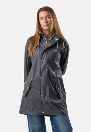 Mujer con un impermeable gris oscuro con bolsillos con botones y cremallera frontal, de pie contra un fondo claro.