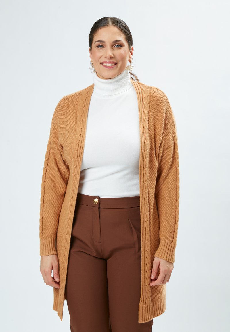 Strikket cardigan i kamelfarve med kabeldetaljer og ribstrikkede манchetter. Båret over en hvid turtleneck og parret med brune høj talje bukser.
