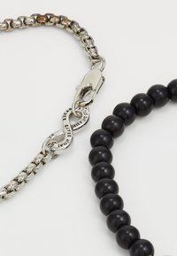 Collana a catena argentata con chiusura a moschettone, abbinata a una collana con perline nere. Presenta un connettore inciso con la scritta "ICON."