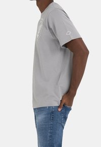 Persona vistiendo una camiseta de manga corta gris claro con un gráfico circular blanco, jeans azules, con una mano en el bolsillo, de lado, sobre un fondo liso.