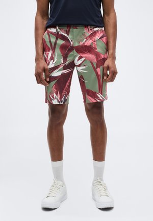 Άνδρας που φοράει κοντά παντελόνια με τροπικό σχέδιο σε πράσινο και βουργουνδί, λευκές κάλτσες, λευκά αθλητικά παπούτσια και μπλε πουκάμισο, στέκεται μπροστά σε απλό φόντο.