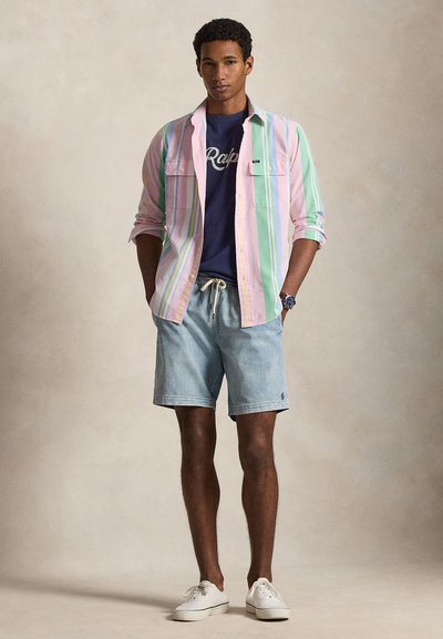 Άνδρας με παστέλ πουά πουκάμισο πάνω από μπλε μπλουζάκι, ανοιχτόχρωμα τζιν σορτς, λευκά αθλητικά παπούτσια και ρολόι, στέκεται με τα χέρια στις τσέπες.