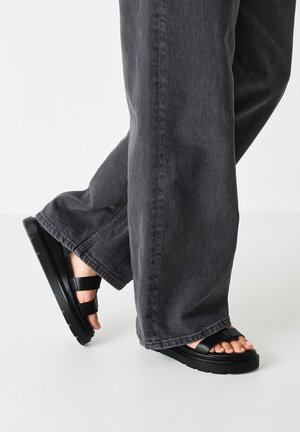 Sandalias de plataforma negras con suela gruesa, dos correas ajustables sobre el pie, combinadas con jeans de pierna ancha grises. Textura suave y diseño minimalista.