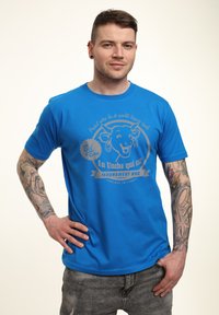 Modrá bavlněná tričko s výraznou krémovou grafikou zobrazující kreslenou krávu a text. Krátké rukávy a kulatý výstřih.