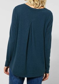 Mörk turkos långärmad topp i mjukt tyg, med en subtil plissér bak och en avslappnad passform med rundad nederkant.