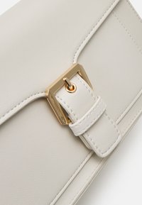 Sac à main en cuir blanc avec un rabat, quincaillerie dorée, une bandoulière et des détails de couture visibles.