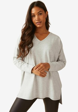Top V-neck à manches longues gris clair avec une coupe décontractée et une fente sur le côté, associé à des leggings noirs. Tissu doux et lisse et design minimal.