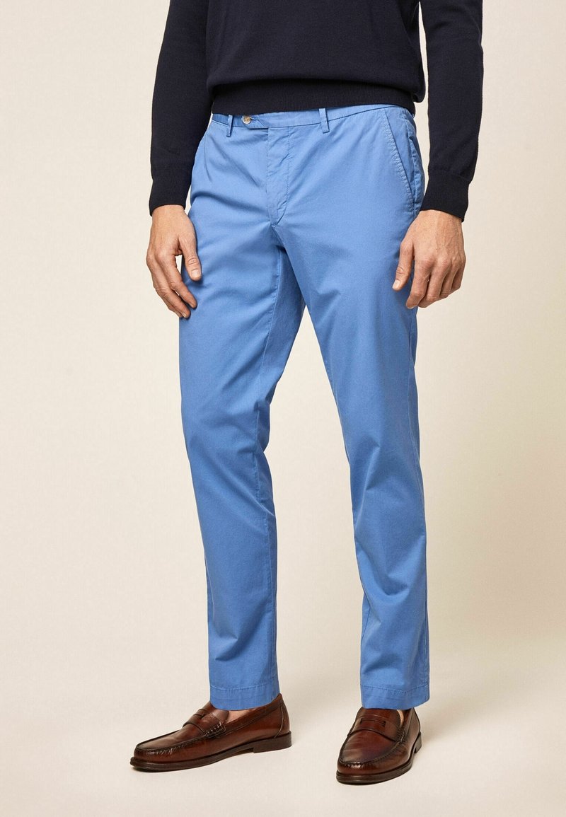 Homme portant un pantalon slim bleu clair, des mocassins en cuir marron et une chemise noire à manches longues, debout devant un arrière-plan neutre.