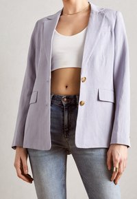 Garnitur o jasnofioletowym kolorze, o fasonie dopasowanym, zapinany na jeden rząd, z dwoma złotymi guzikami i kieszeniami z patkami, zestawiony z niebieskimi dżinsami.