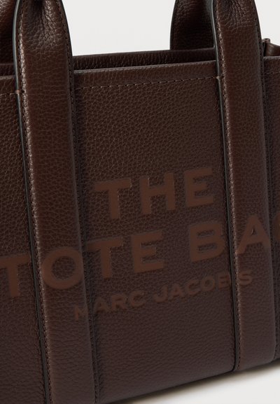 Τσάντα tote από σκοτεινό καφέ δέρμα με υφή, διπλές λαβές και ανάγλυφο κείμενο «THE TOTE BAG MARC JACOBS» στο πλάι.