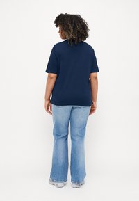 Maglietta in cotone blu navy con maniche corte e vestibilità rilassata, indossata con jeans a zampa d'elefante blu chiaro e sneakers grigie, vista da dietro.