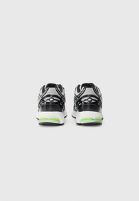 Paire de chaussures de sport noires, grises et blanches avec des touches vertes, vues de dos sur un fond gris clair uni.