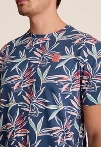 Marineblauwe t-shirt met een tropisch bladprint in groen, rood en roze. Heeft een klein oranje logo-opdruk op de borst. Korte mouwen.