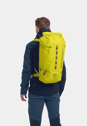 Bright yellow Ortovox backpack with a roll-top design, external straps, and logo. Worn by a person dressed in dark outerwear and pants.