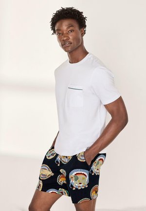 Junger Mann trägt ein weißes T-Shirt mit Tasche und schwarze Shorts mit bunten Elefanten- und Tiermotiven, Hände in den Taschen, neutraler Hintergrund.