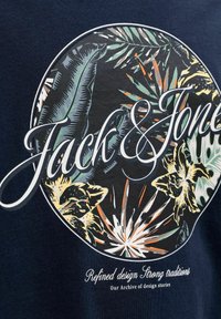 Motif circulaire floral avec le grand texte « Jack & Jones » sur des feuilles tropicales et des fleurs sur tissu foncé, avec un texte plus petit en dessous.
