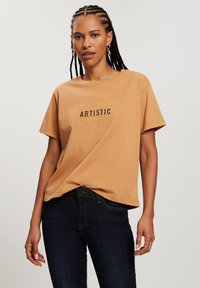 Beżowa bawełniana koszulka z krótkim rękawem i prostokątnym nadrukiem z napisem "ARTISTIC" w kolorze czarnym. W połączeniu z ciemnoniebieskimi dżinsami.