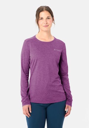Donna con maglia a maniche lunghe viola con logo Vaude sul petto e pantaloni blu scuro, in piedi contro uno sfondo chiaro e uniforme.
